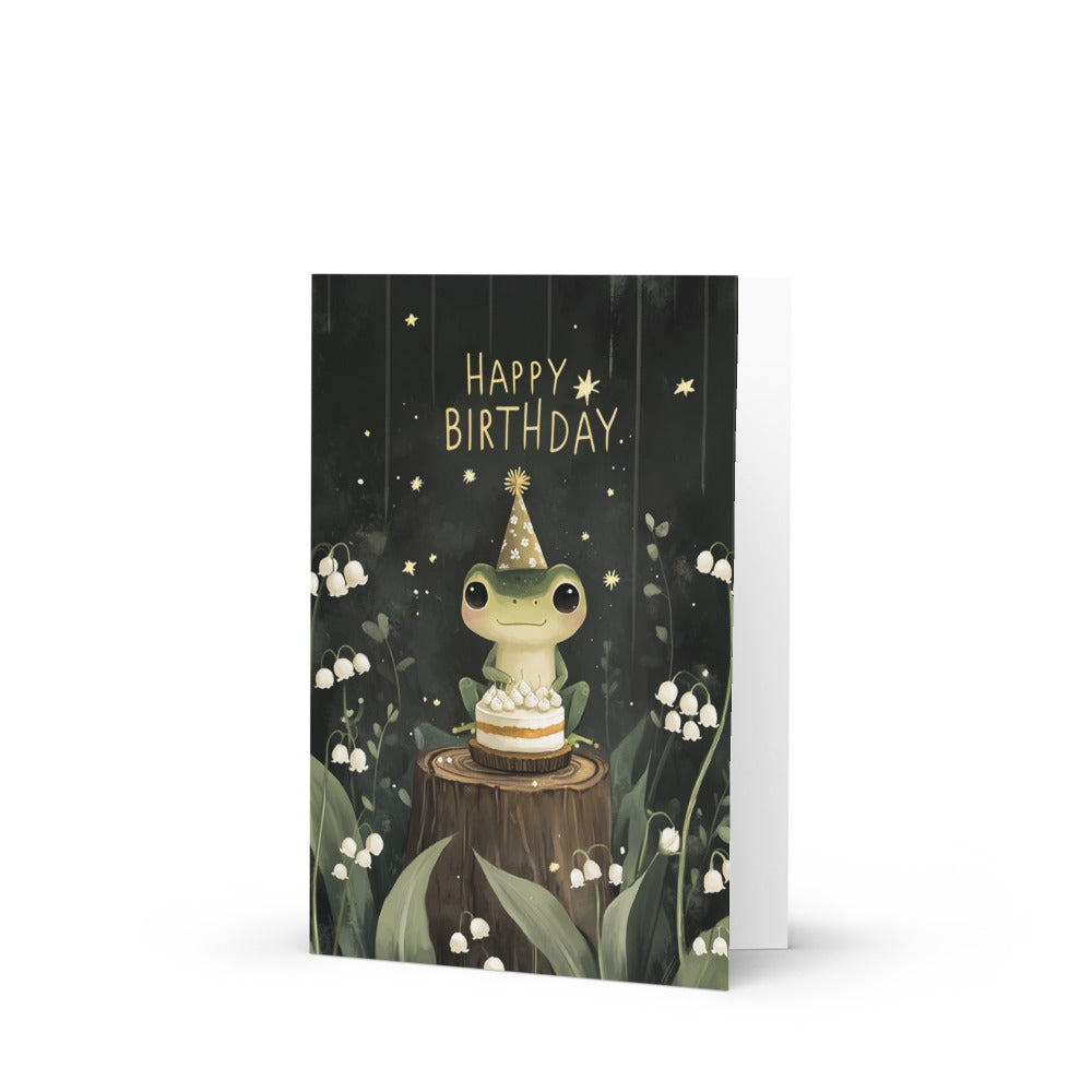 A whimsical Kawaii frog birthday card featuring a cute frog wearing a party hat, sitting on a tree stump with a slice of cake, surrounded by white lilies and lush greenery, perfect for frog lovers and birthday celebrations.